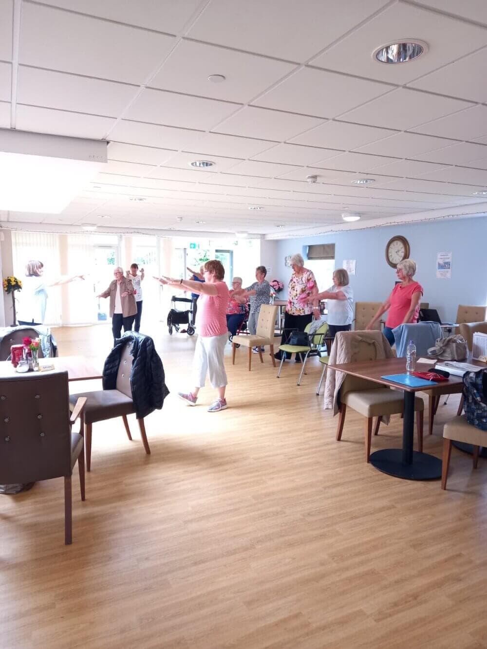 Group of elderly people participating in a fitness class in a room with wooden flooring and tables. - Home Instead