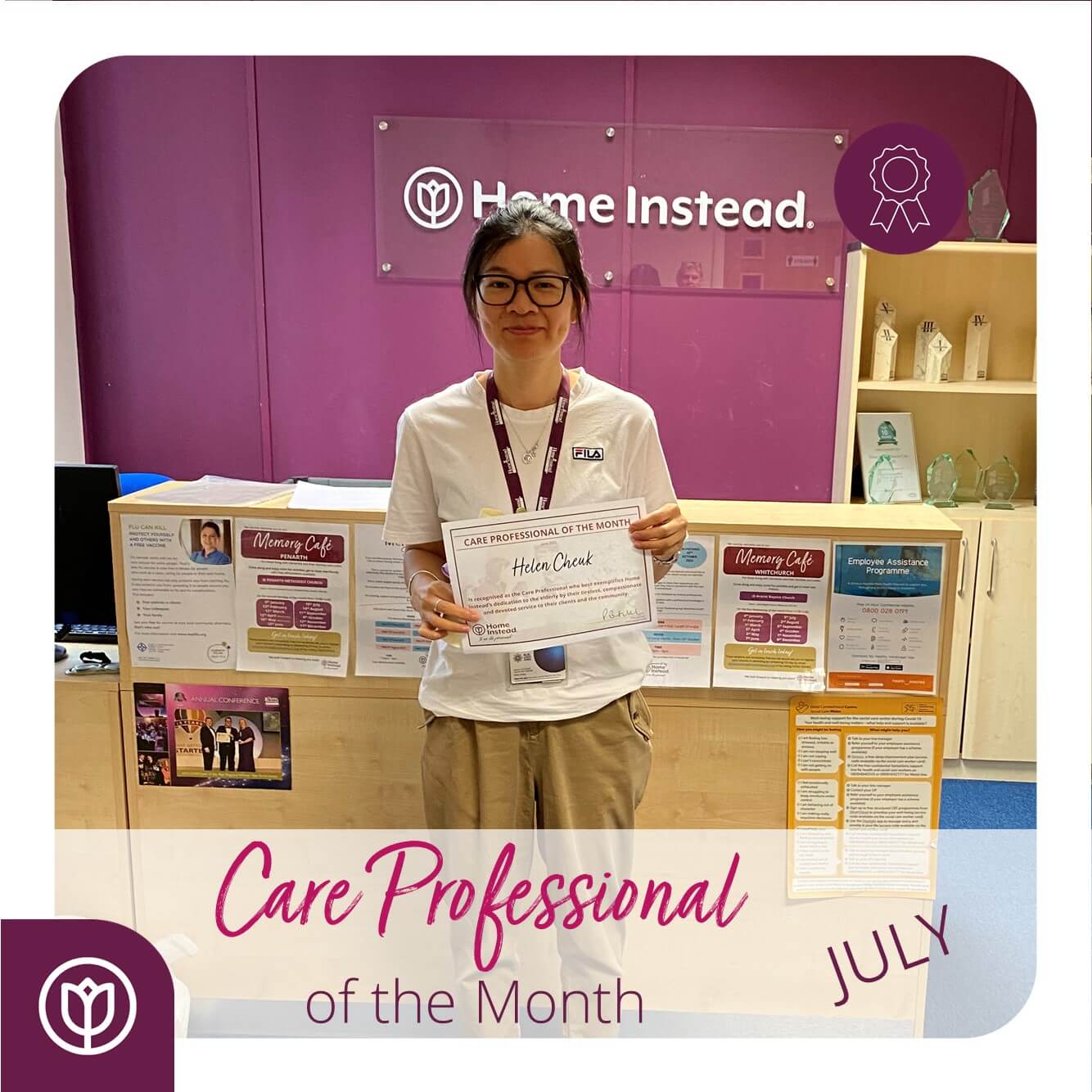 Person holding a "Care Professional of the Month" certificate, standing in front of a "Home Instead" office display. - Home Instead