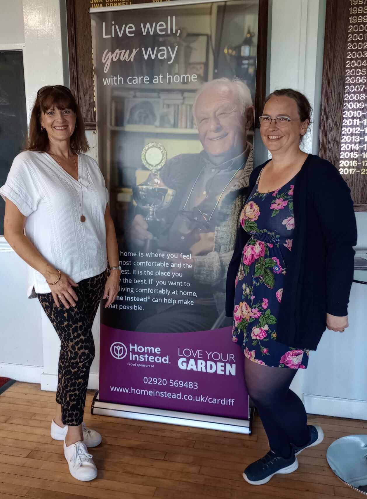 Two women standing beside a banner for Home Instead's care services, smiling at the camera. - Home Instead