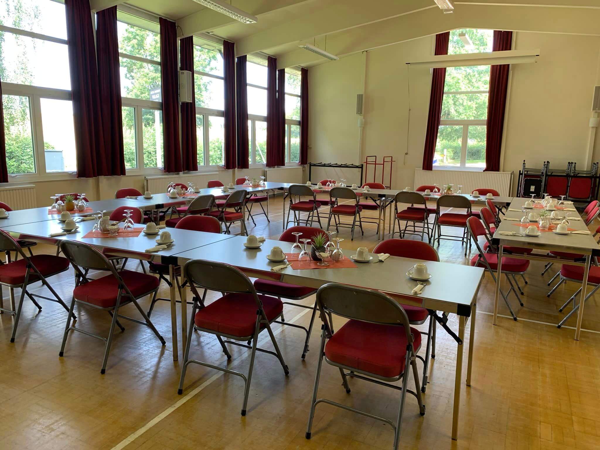A hall set up with tables, chairs, and place settings, ready for an event, with natural light streaming through large windows. - Home Instead
