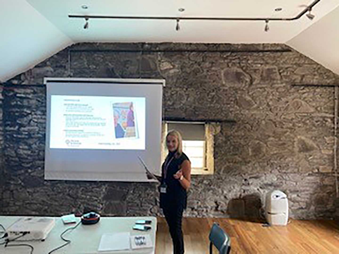 A woman stands near a projector screen giving a presentation in a room with stone walls and a wooden floor. - Home Instead