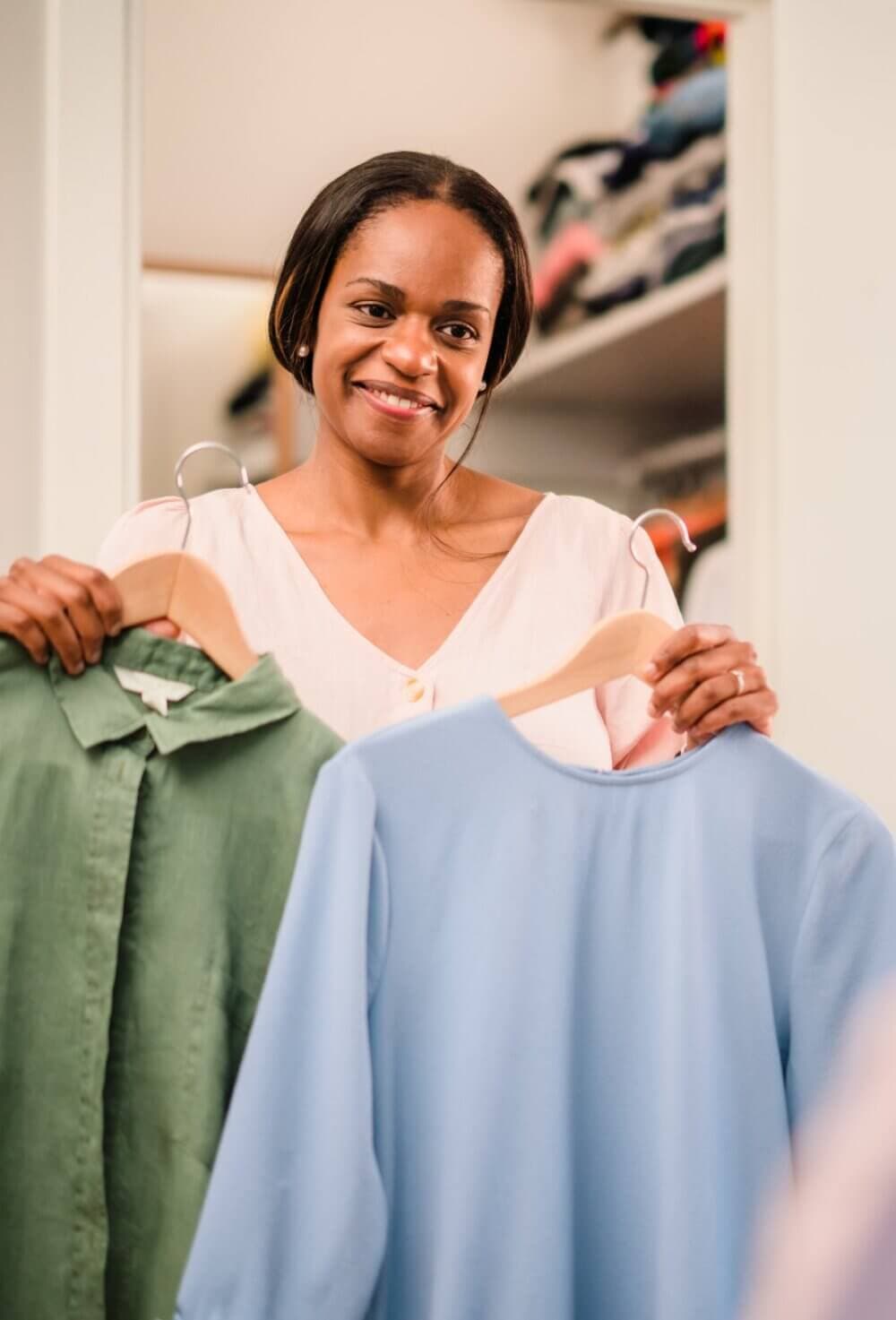 A woman smiles, holding a green shirt and a blue shirt on hangers in front of her in a walk-in closet. - Home Instead