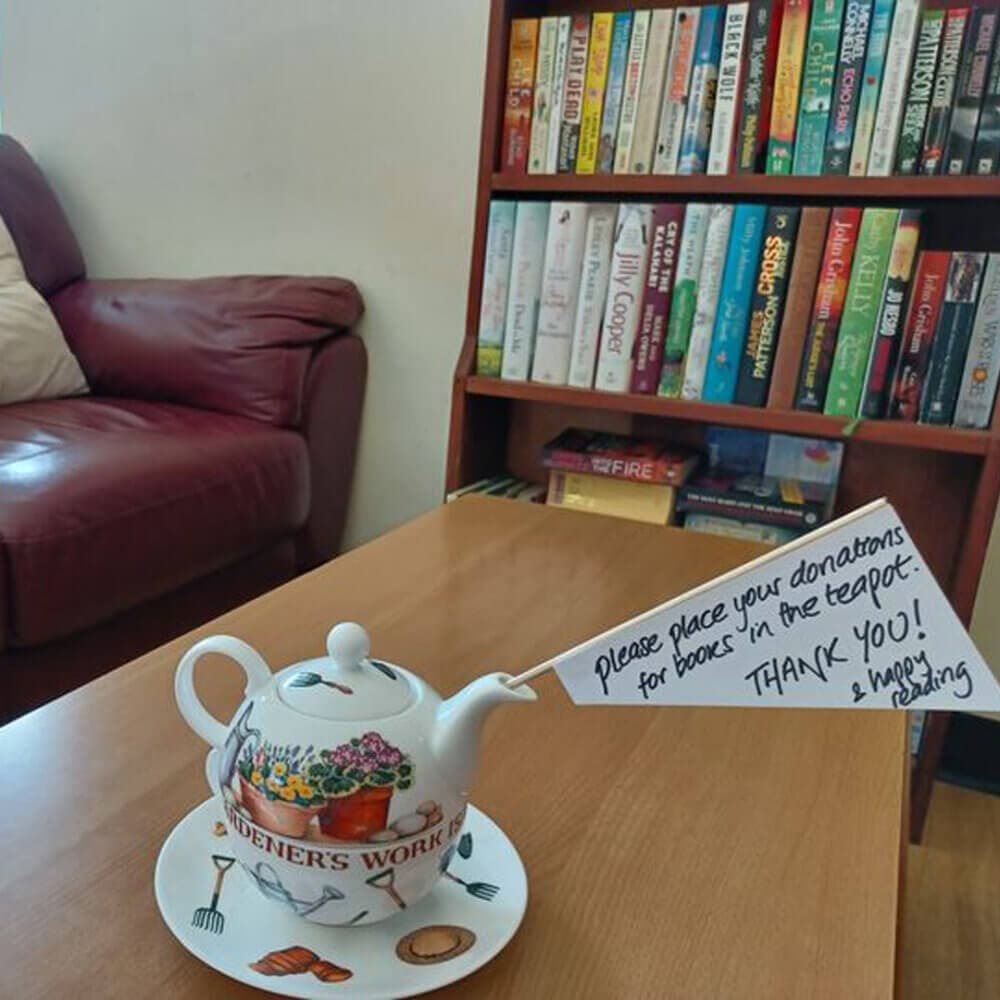 A teapot with a note requesting book donations sits on a table in a cozy room with a bookshelf and couch. - Home Instead