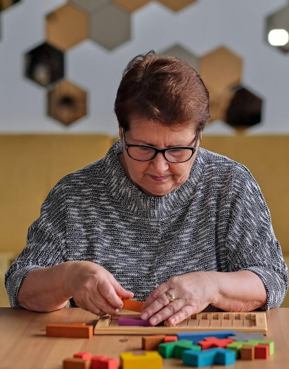 A woman with glasses focuses on assembling a colorful geometric puzzle at a table. - Home Instead