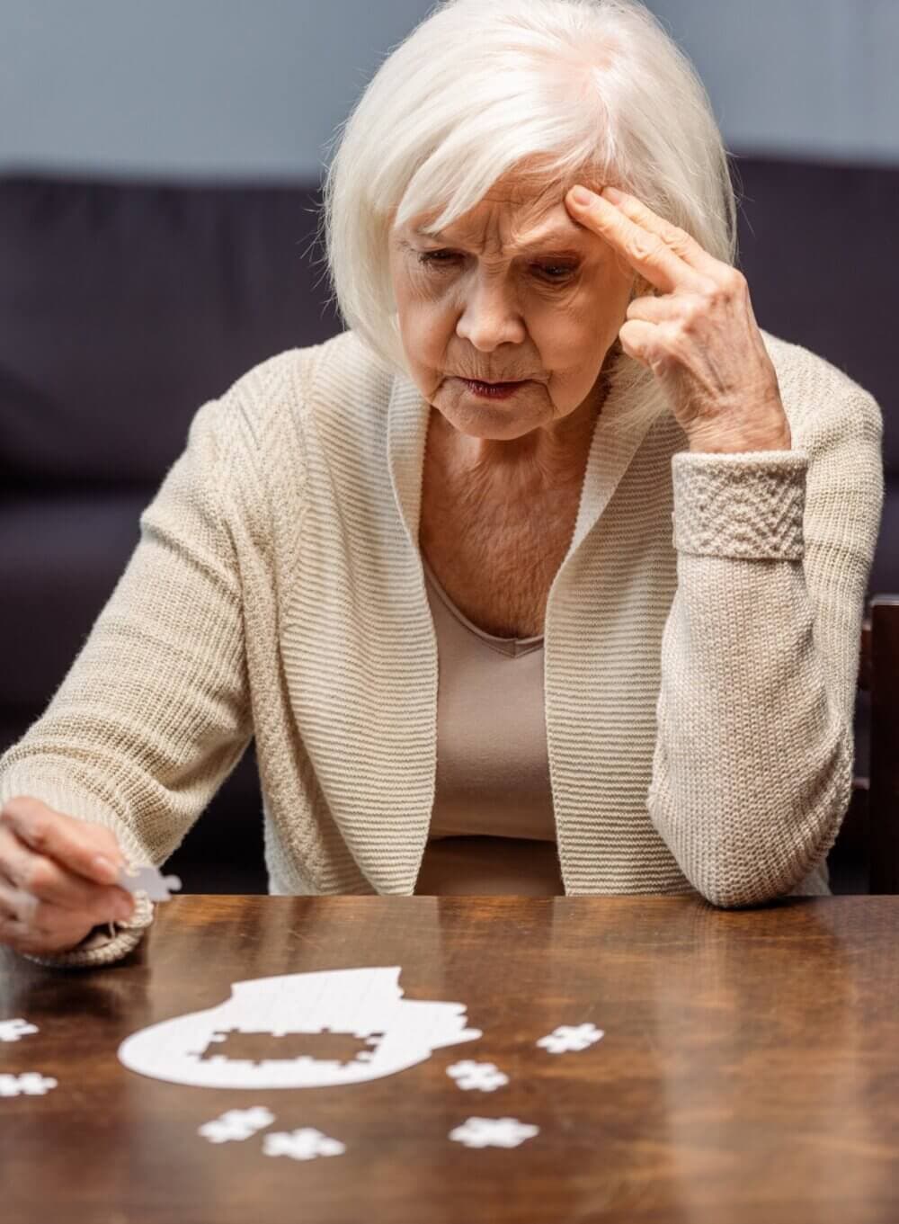 An elderly woman in a beige cardigan concentrates on solving a jigsaw puzzle at a table. - Home Instead
