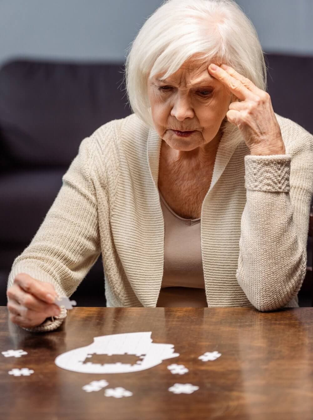 An elderly woman with white hair looks concerned while solving a puzzle at a wooden table. - Home Instead