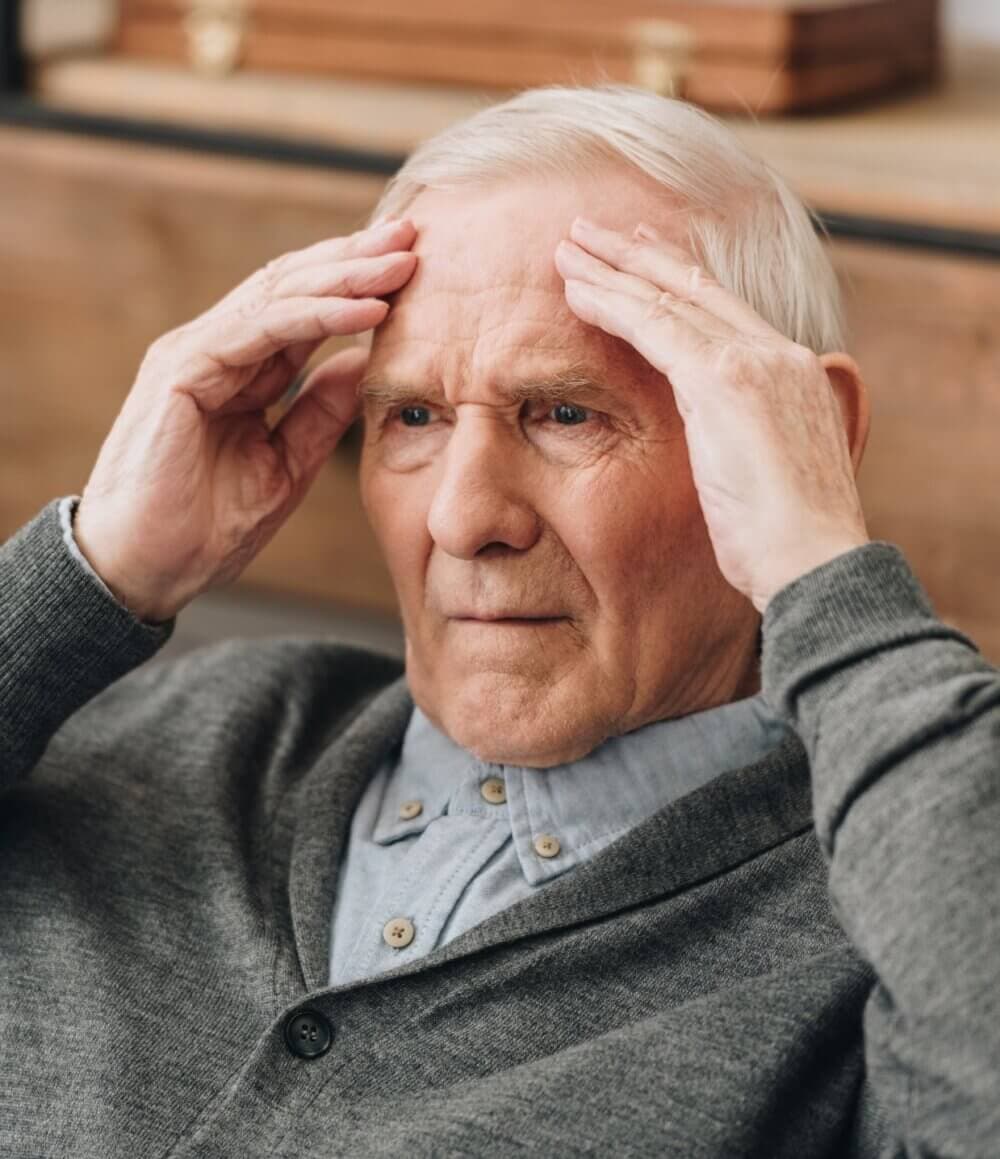 Elderly man in gray cardigan with hands on head, appearing stressed or deep in thought, indoors. - Home Instead