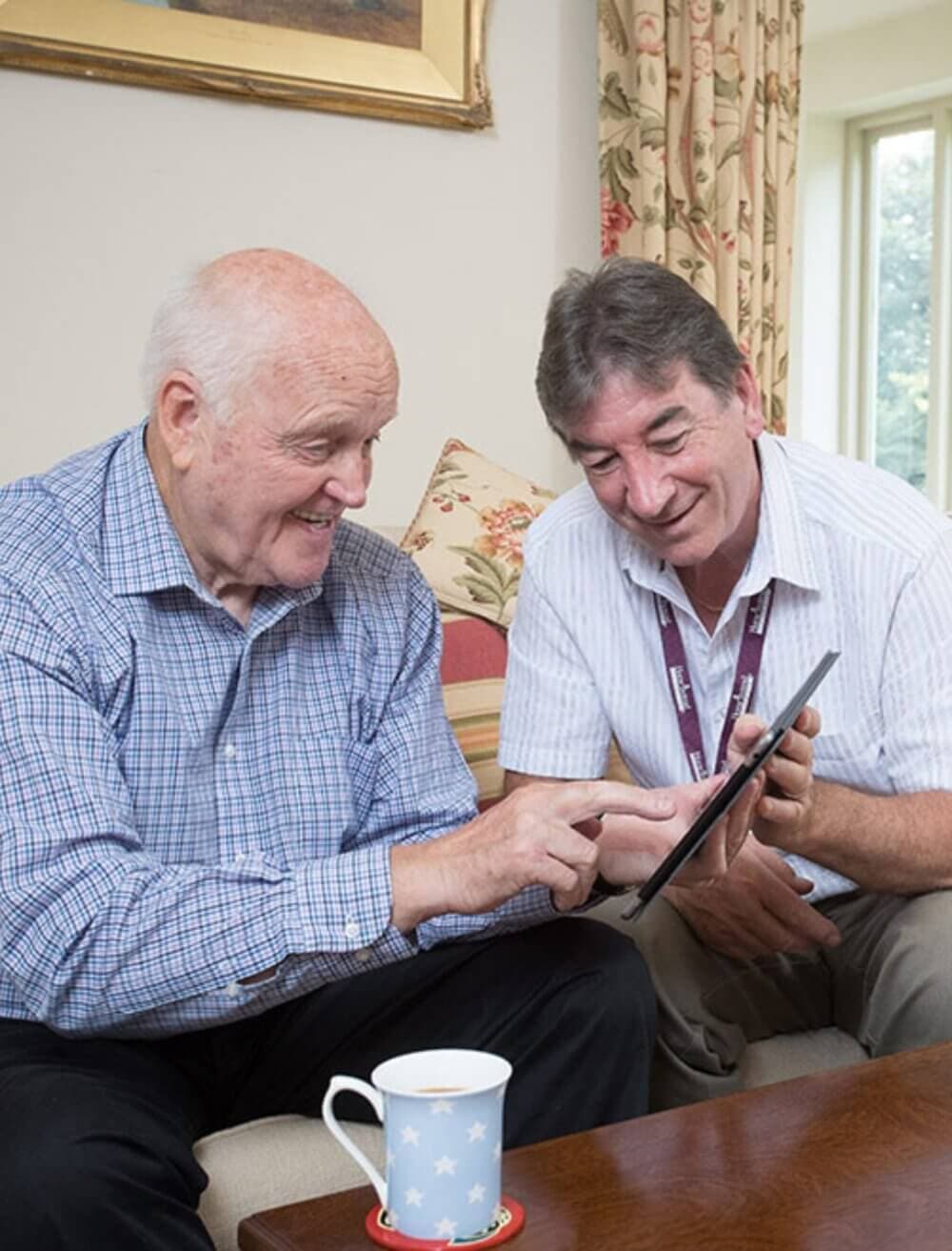 Two men smiling while looking at a tablet, seated on a couch with a coffee mug on a table in front of them. - Home Instead