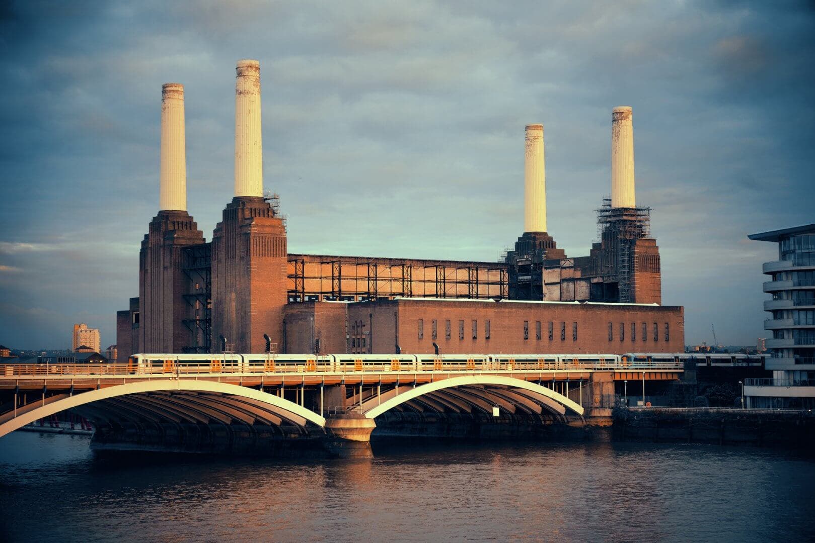A historic industrial building with four tall chimneys near a river, featuring a bridge in the foreground. - Home Instead