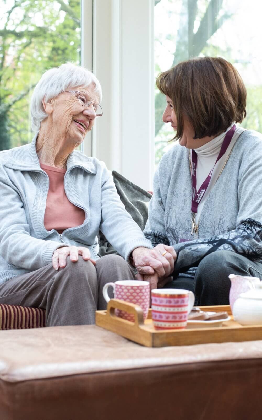 An elderly woman and a younger woman sit on a couch, smiling and holding hands, with tea cups on a tray in front of them. - Home Instead