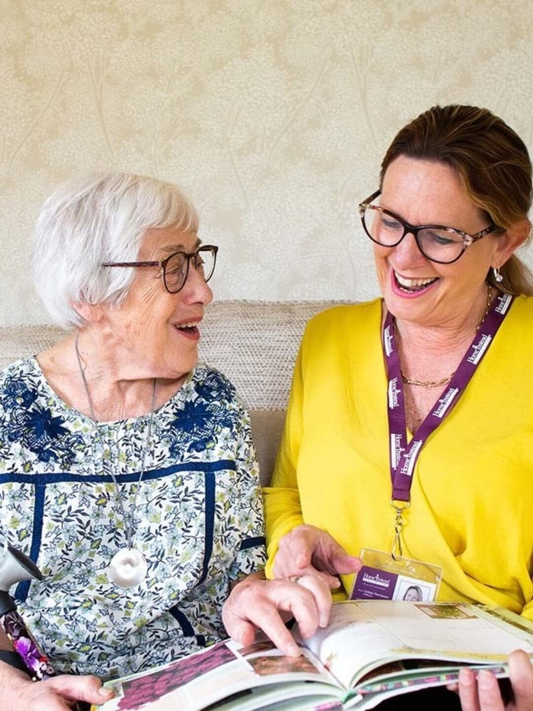 An elderly woman and a caregiver with a lanyard smile and look at an open book together. - Home Instead