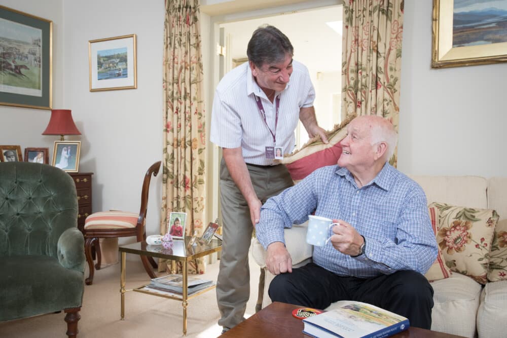 A caregiver stands smiling over an elderly man who is sitting and holding a mug in a cozy living room. - Home Instead