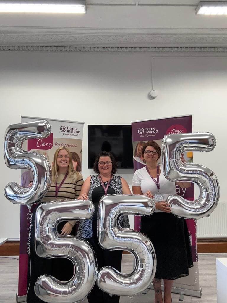 Three people smiling and holding large silver balloon numbers "555" indoors, with banners in the background. - Home Instead