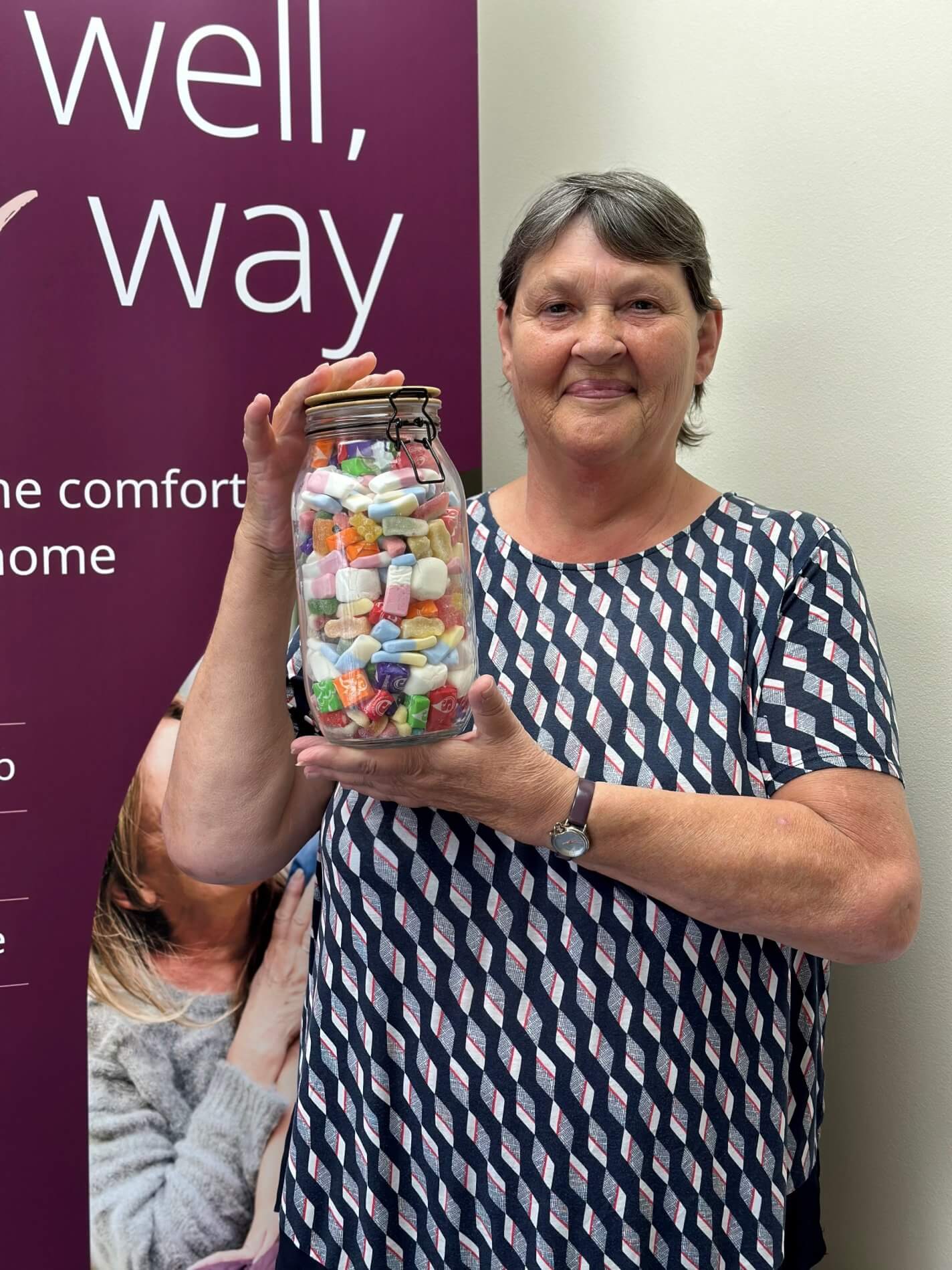 A woman holding a large jar of colorful candies, standing in front of a banner with partial text visible. - Home Instead
