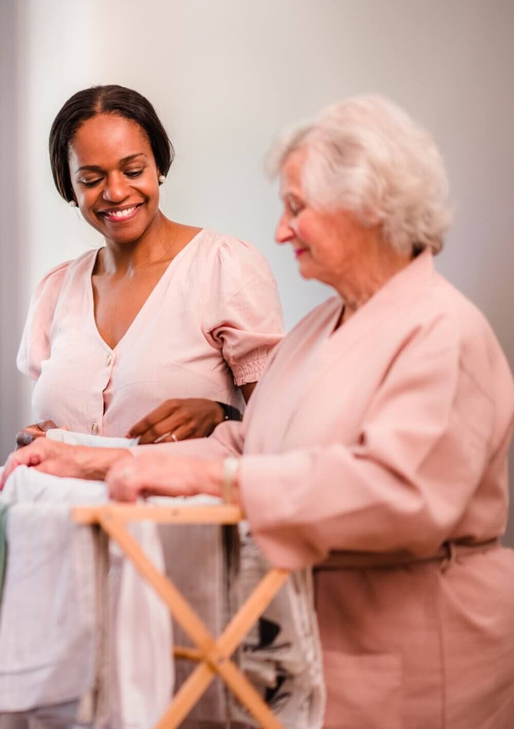 A smiling woman helps an elderly woman fold laundry on a wooden drying rack. - Home Instead
