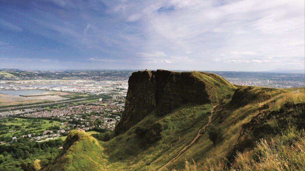 Aerial view of a grassy hill with cliffs overlooking a city, a river, and distant landscape under a partly cloudy sky. - Home Instead