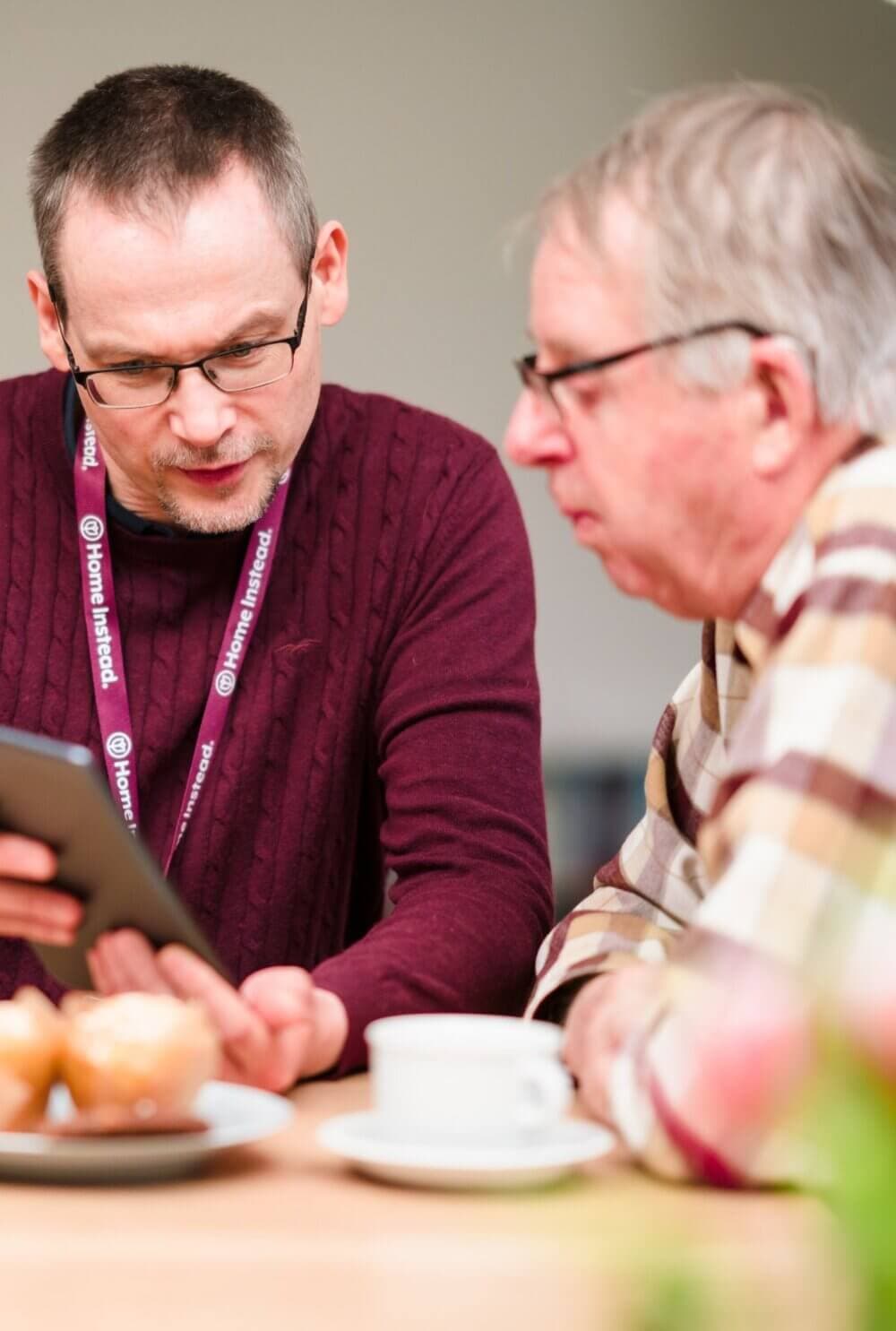Two men, one holding a tablet, sit at a table with a coffee cup and muffins, engaged in conversation. - Home Instead