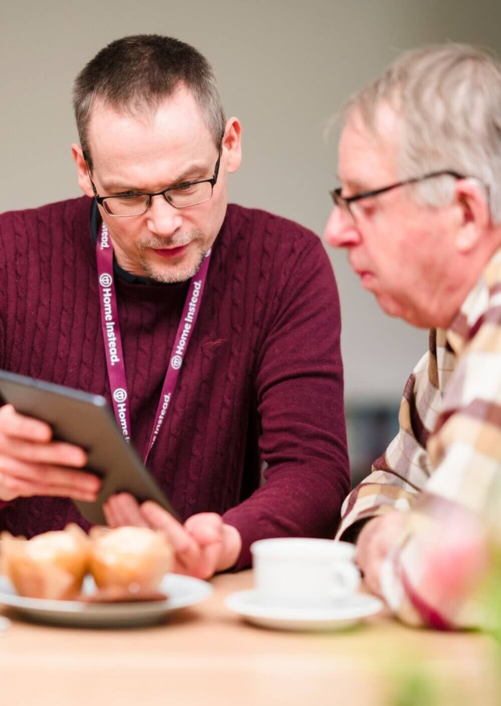 Two men sitting at a table with muffins and a cup of coffee, looking at a tablet together. - Home Instead