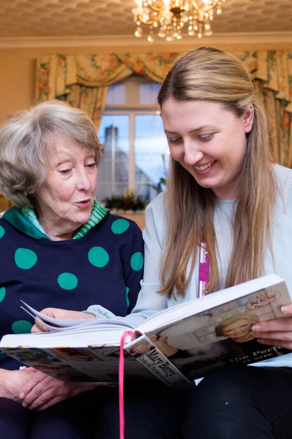 An elderly woman and a younger woman smiling while looking at a large book together in a cozy room. - Home Instead