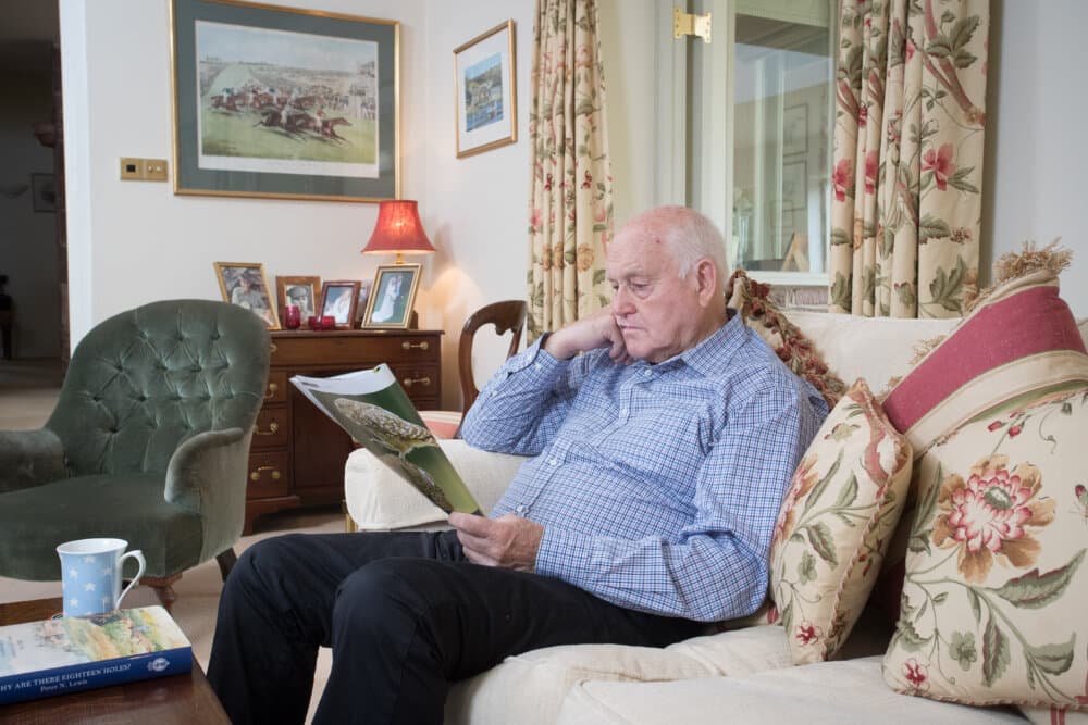 An elderly man reads a magazine in a cozy living room with floral curtains and framed wall art. - Home Instead
