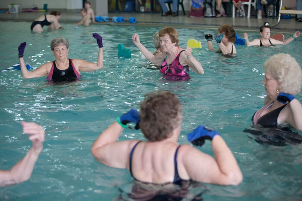 Older adults participating in an aquatic exercise class in an indoor swimming pool, wearing swimwear and gloves. - Home Instead