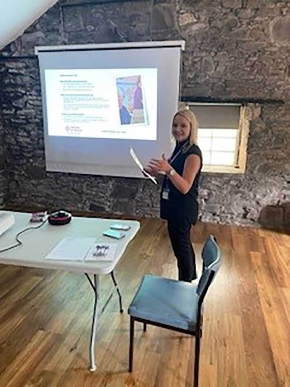 Person giving a presentation in a room with a stone wall, a projector screen, and a table with materials on it. - Home Instead