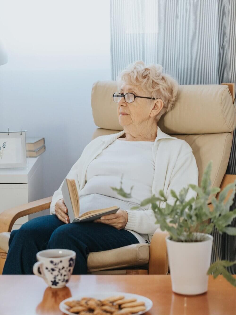 Elderly woman with glasses reading a book while sitting on a chair next to a table with a plant, cup, and plate of cookies. - Home Instead Bournemouth & Christchurch