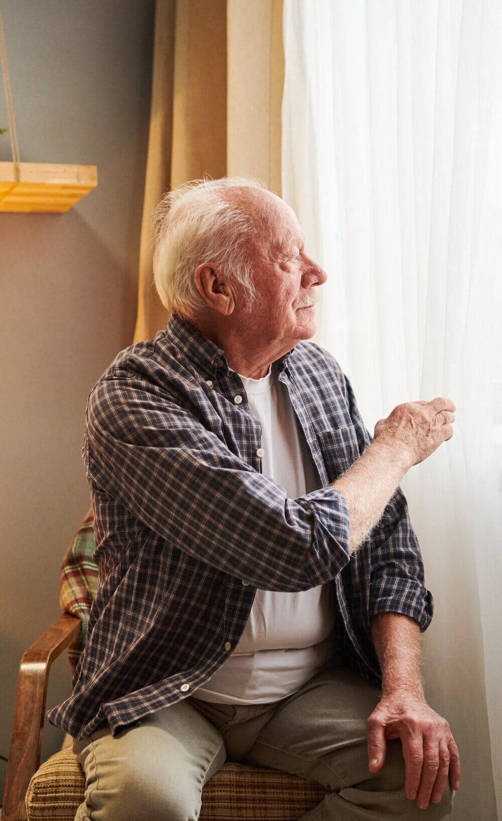 Elderly man with white hair and plaid shirt looks out of a window, pulling back the curtain. - Home Instead