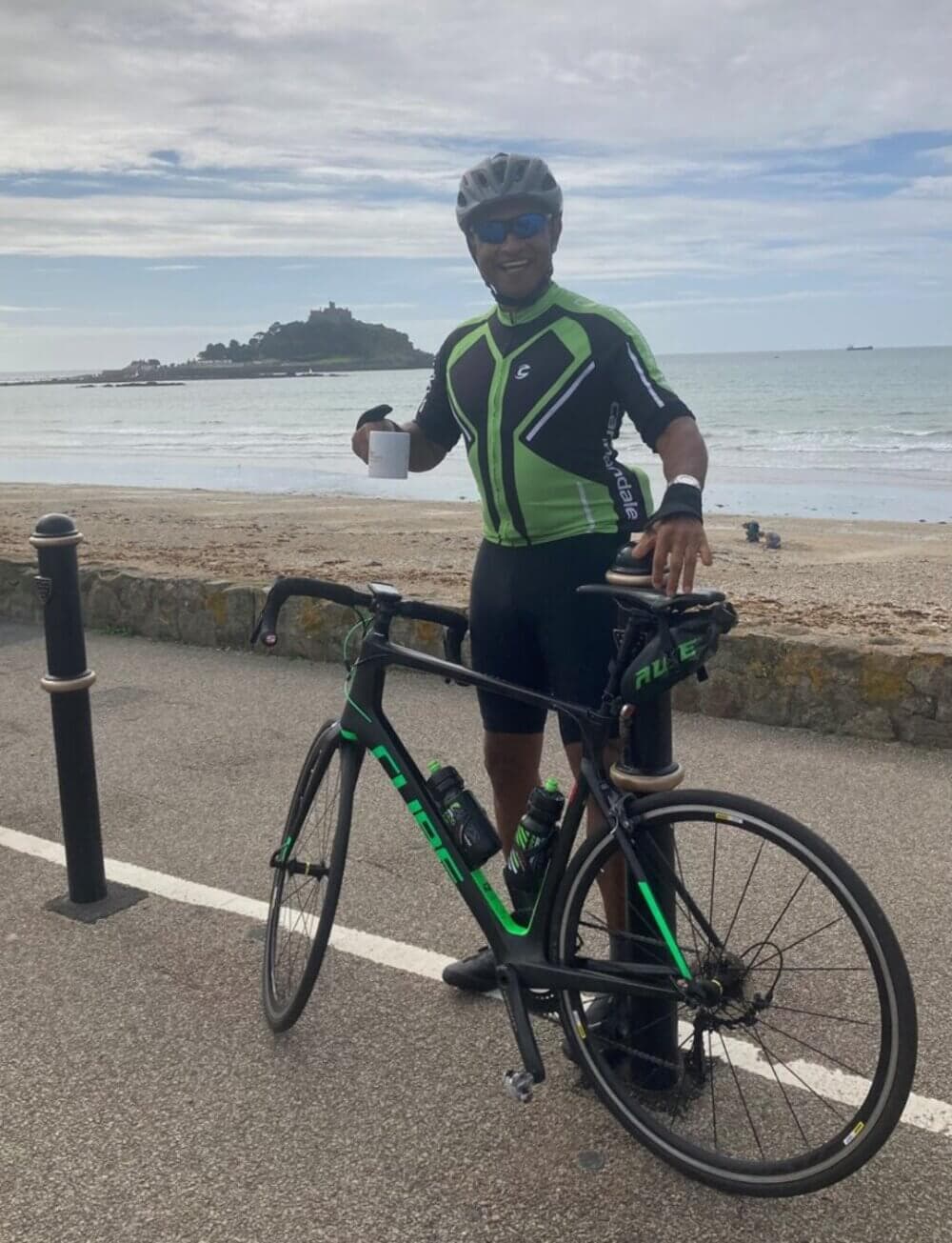 Cyclist in green and black gear holds a mug beside a bike, with a beach and an island in the background. - Home Instead