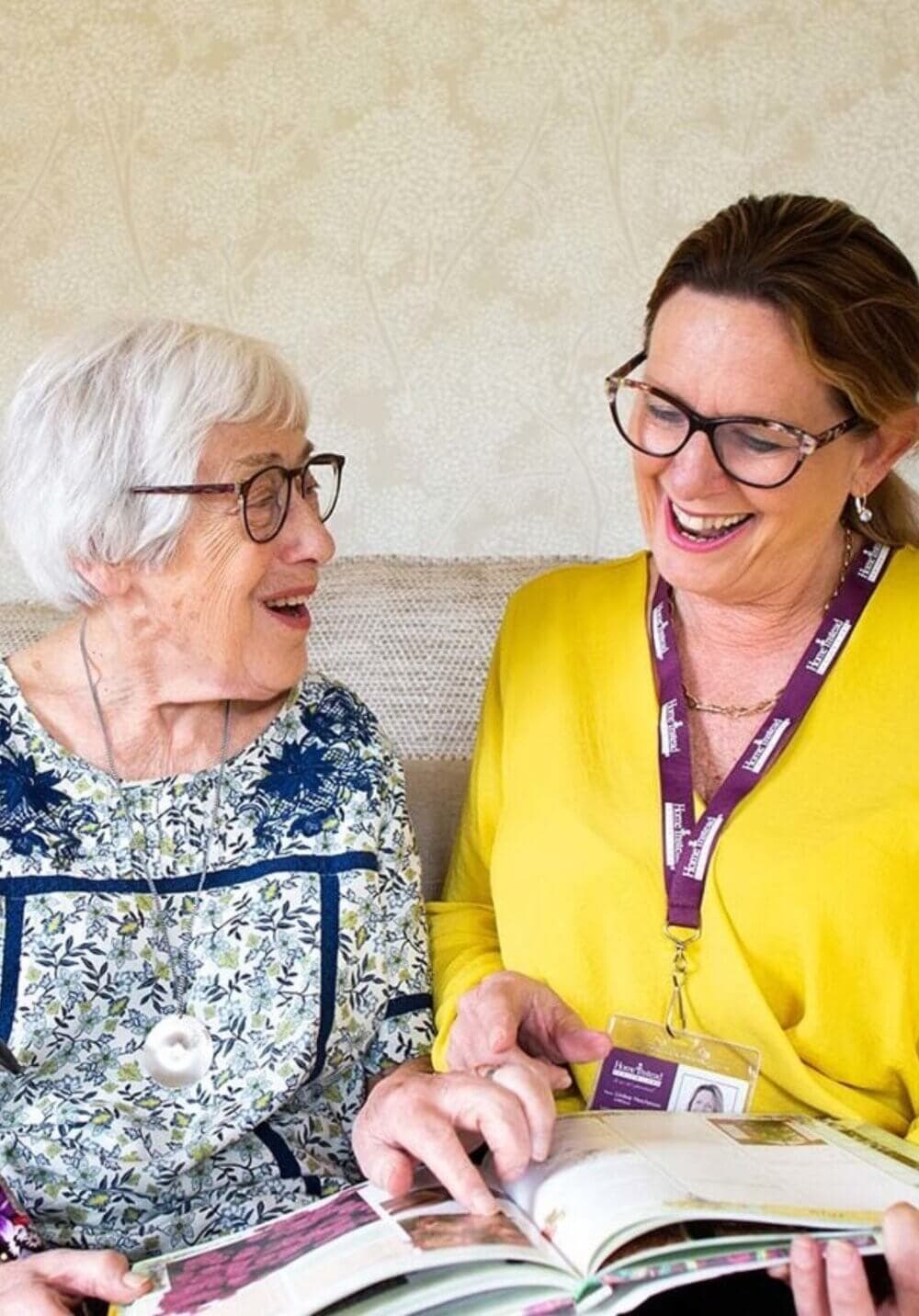 Elderly woman and caregiver in yellow shirt and glasses smiling and looking at a book together. - Home Instead