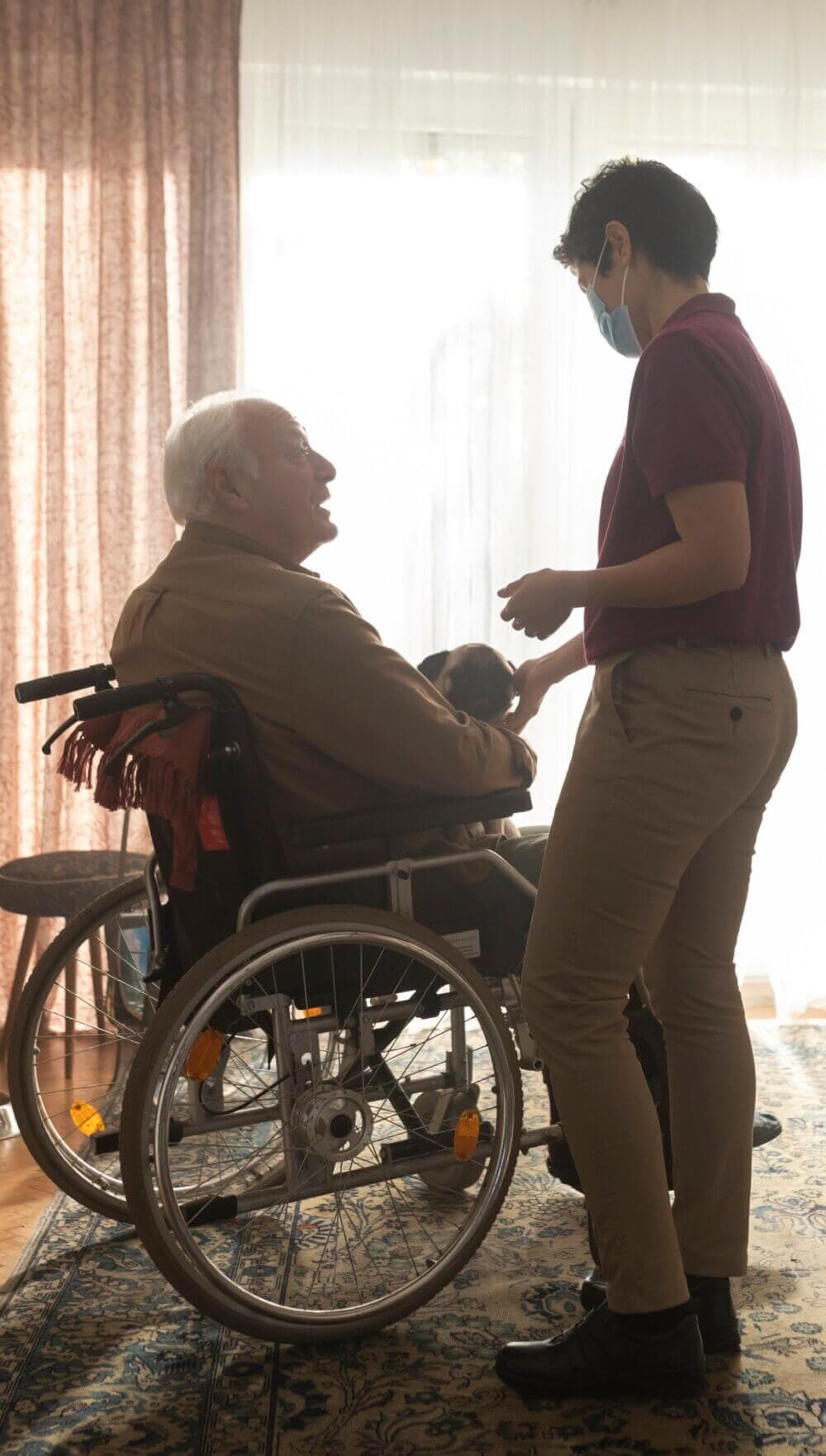 A young caregiver in a mask holds hands with an elderly man in a wheelchair inside a bright room with large windows. - Home Instead