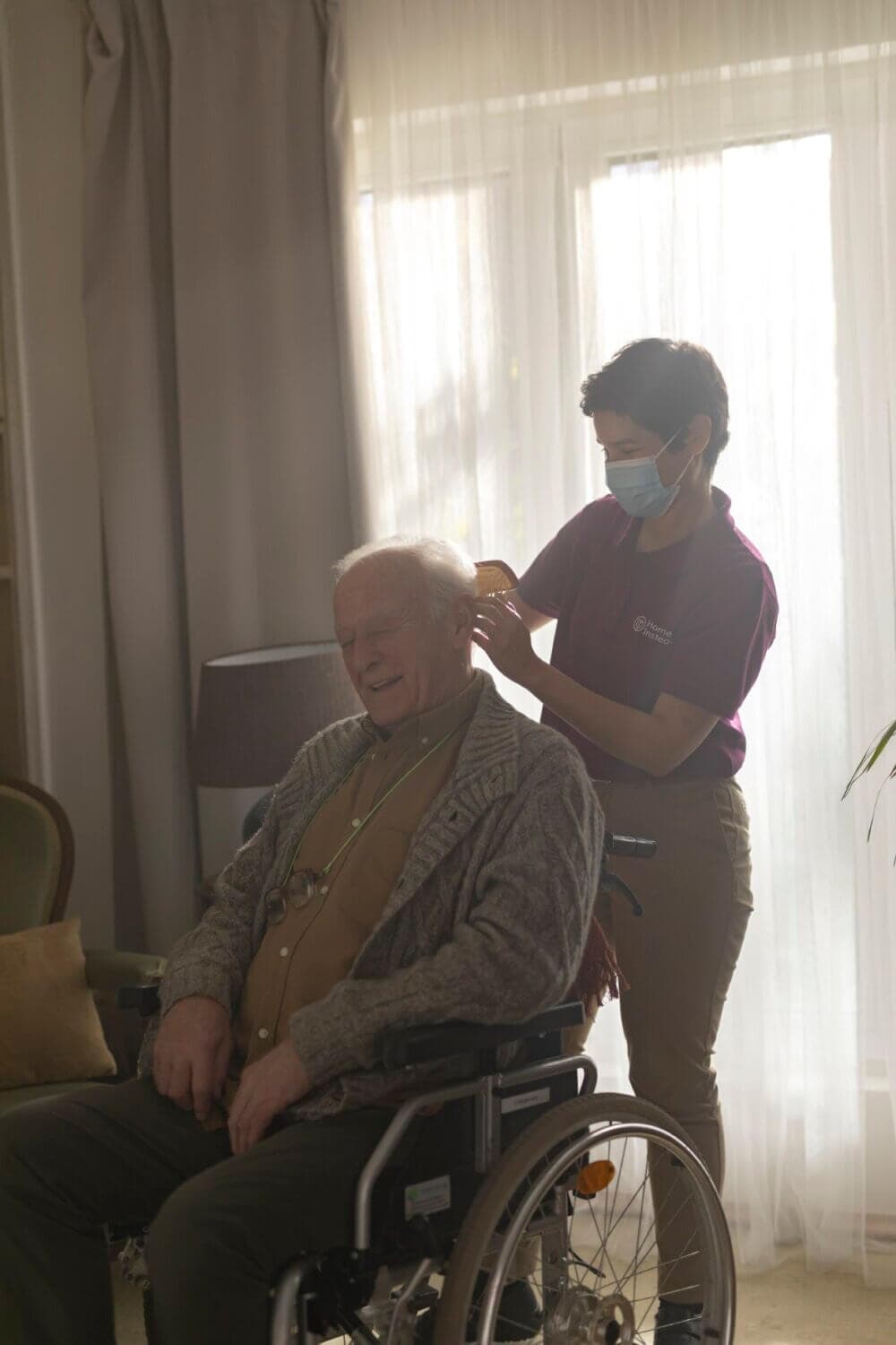 A caregiver puts a mask on a smiling elderly man in a wheelchair, with sunlight streaming through a window behind them. - Home Instead