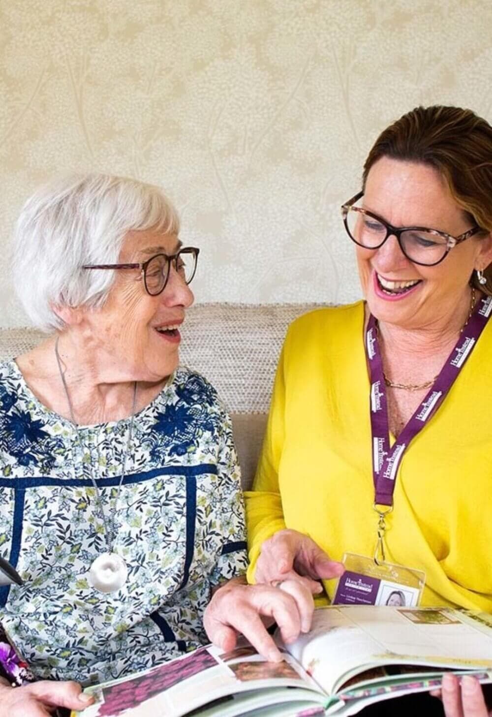 Two women with glasses smiling and reading a book together; one is elderly in floral attire, the other in yellow with an ID badge. - Home Instead Poole
