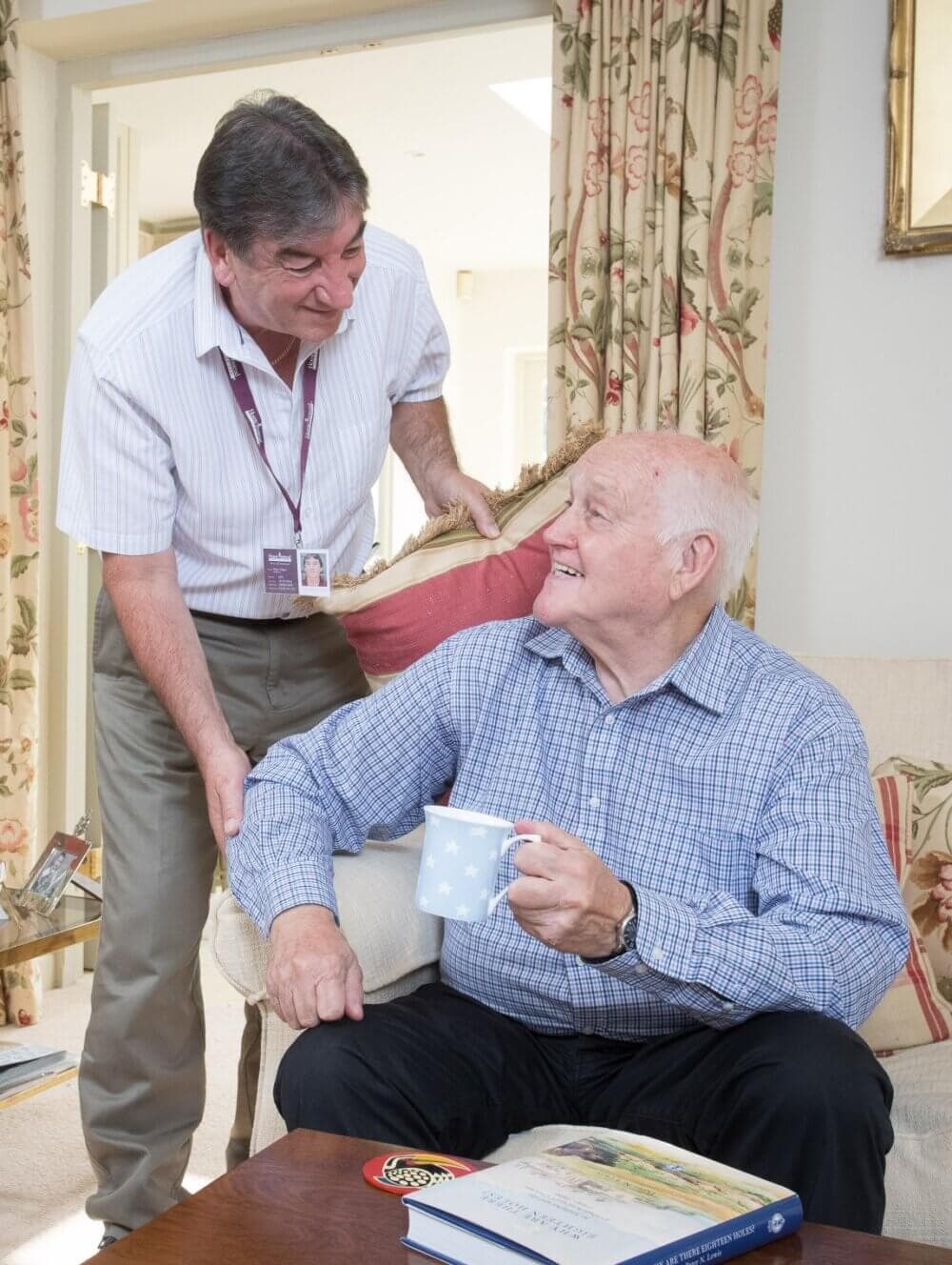 A caregiver smiles and talks with an elderly man holding a mug, in a cozy living room with floral curtains. - Home Instead