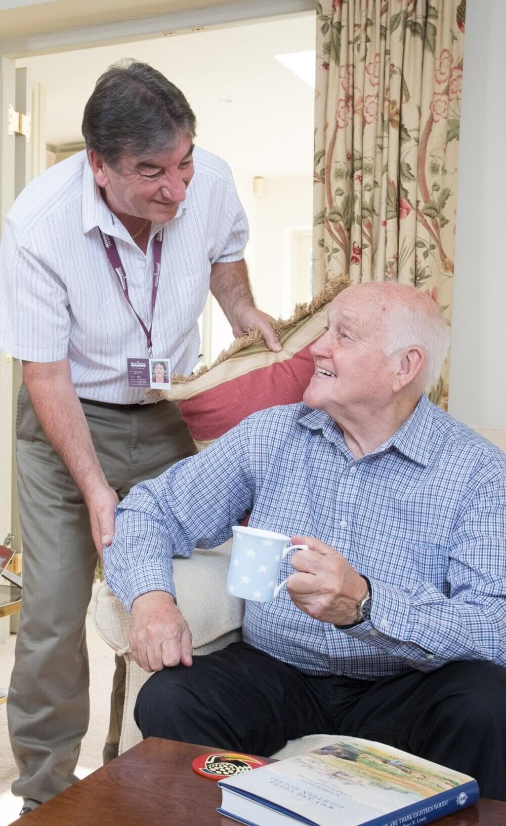 Older man holding a mug and smiling at a younger man who is standing next to him, in a cozy living room. - Home Instead