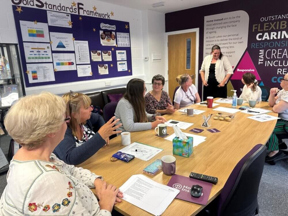 Group of people engaged in a discussion around a conference table in an office setting, with posters and charts on the wall. - Home Instead