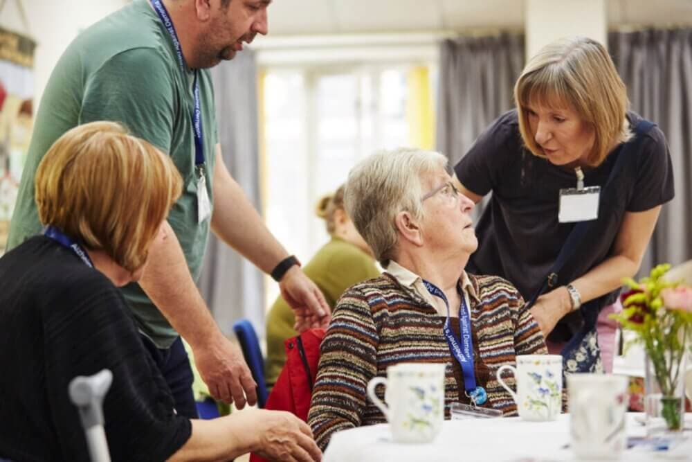 A group of people engaging in conversation around a table with teacups, one woman is seated and others are standing. - Home Instead