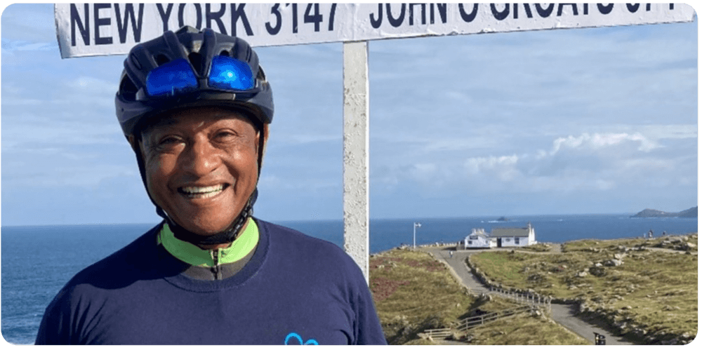 A smiling cyclist wearing a helmet stands in front of a signpost near the coast, under a partly cloudy sky. - Home Instead