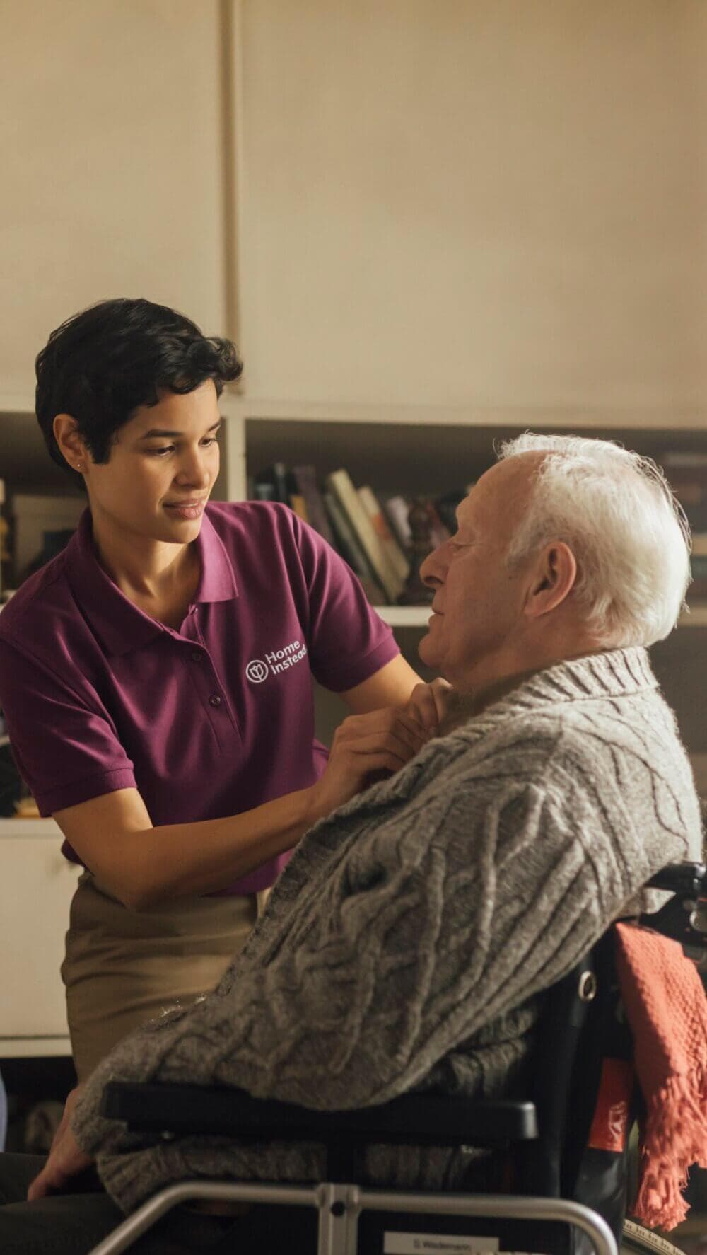 A caregiver in a purple shirt assists an elderly man in a wheelchair with his sweater inside a cozy room. - Home Instead