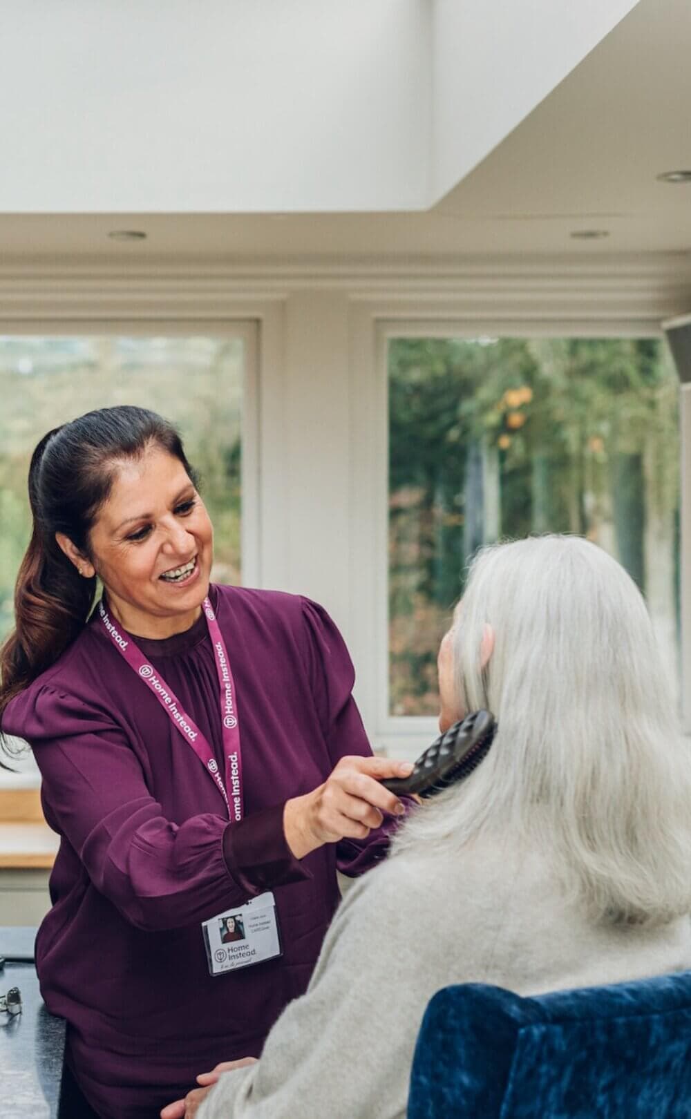 A caregiver in a purple shirt brushes an elderly woman's hair in a bright room with large windows. - Home Instead Southampton