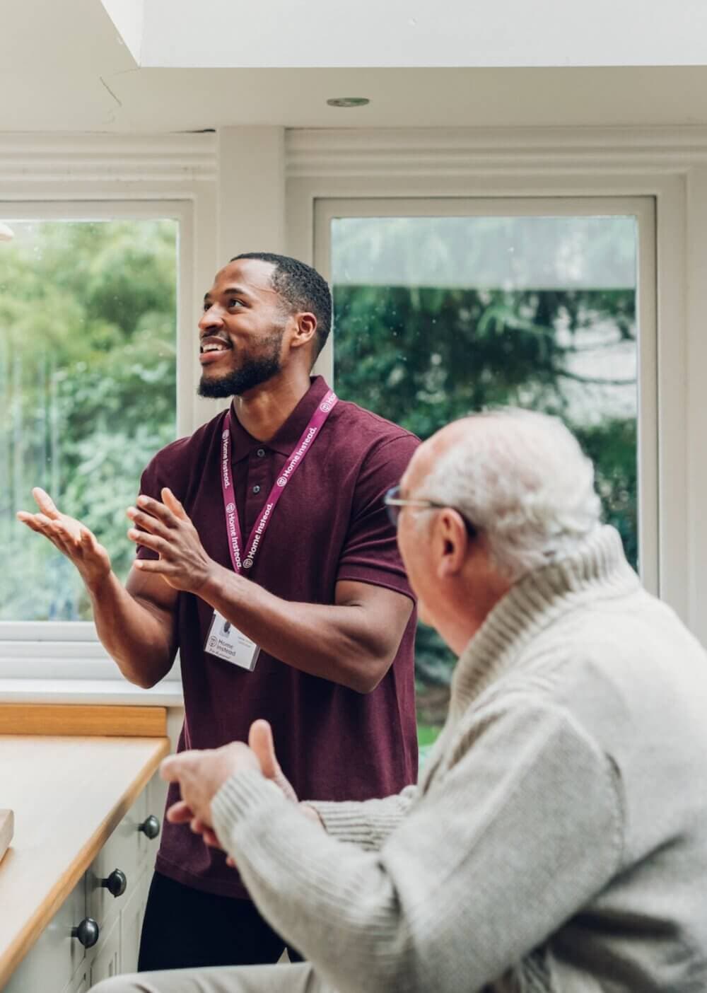 A caregiver in a maroon polo shirt converses with an elderly man in a white sweater in a bright room with large windows. - Home Instead