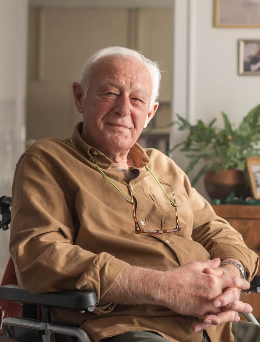 Elderly man with white hair and a brown shirt, sitting in a chair, smiling with glasses hanging around his neck. - Home Instead