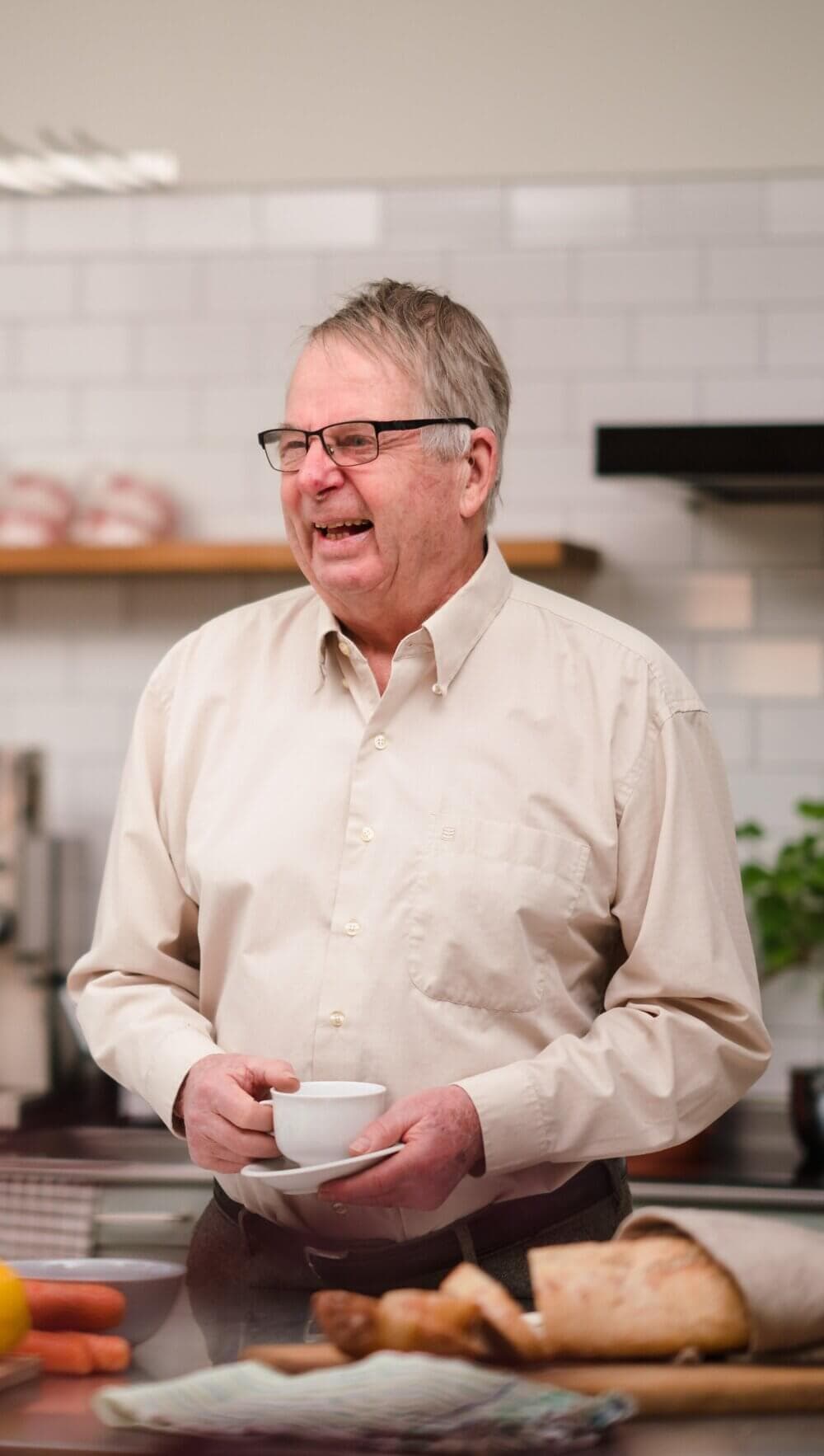 Older man with glasses holding a cup and saucer, smiling in a kitchen with bread and vegetables on the counter. - Home Instead Bournemouth & Christchurch