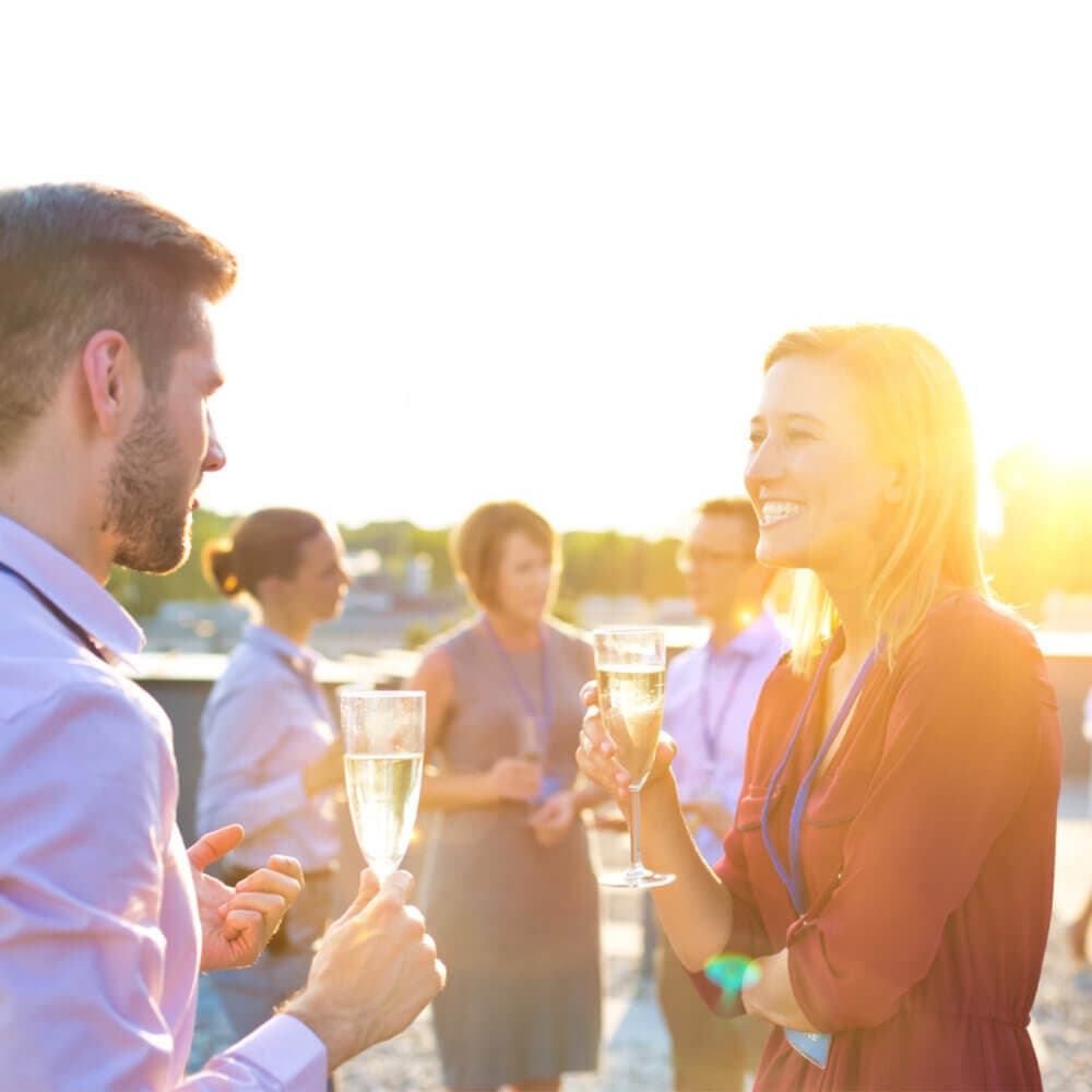 A group of people smiling and holding champagne glasses at an outdoor event during sunset. - Home Instead