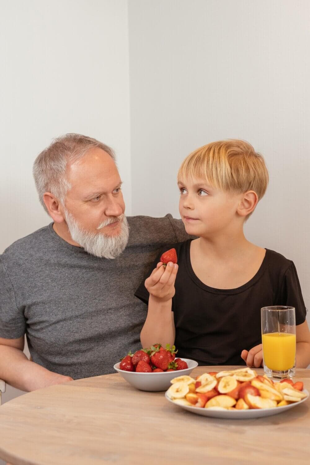 An older man and a child at a table with strawberries, sliced fruit, and a glass of orange juice. - Home Instead