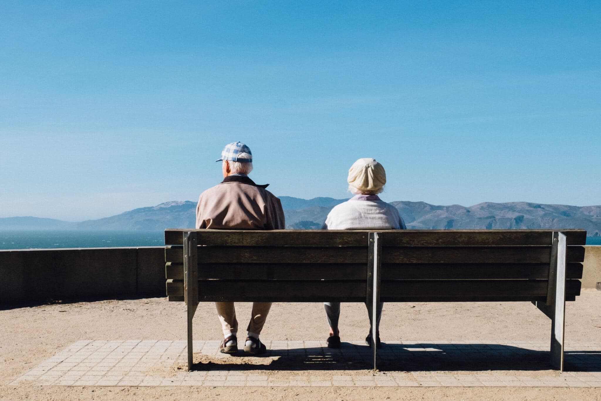 Two elderly people sit on a bench facing a scenic mountain view under a clear blue sky. - Home Instead