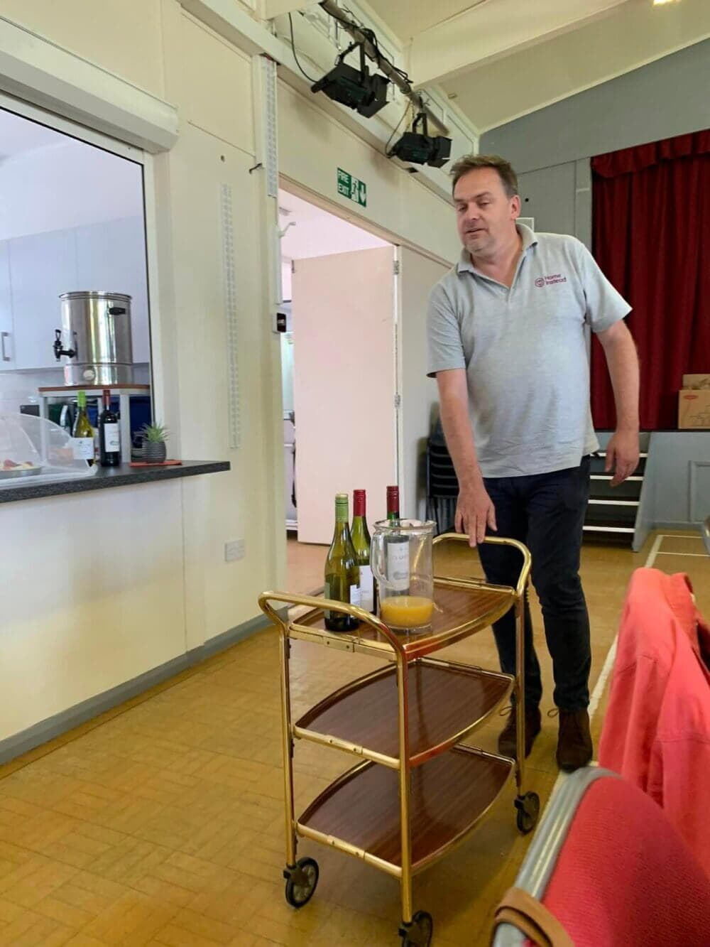 A man rolls a cart with wine bottles and orange juice next to a bar area in a room with a red curtain. - Home Instead