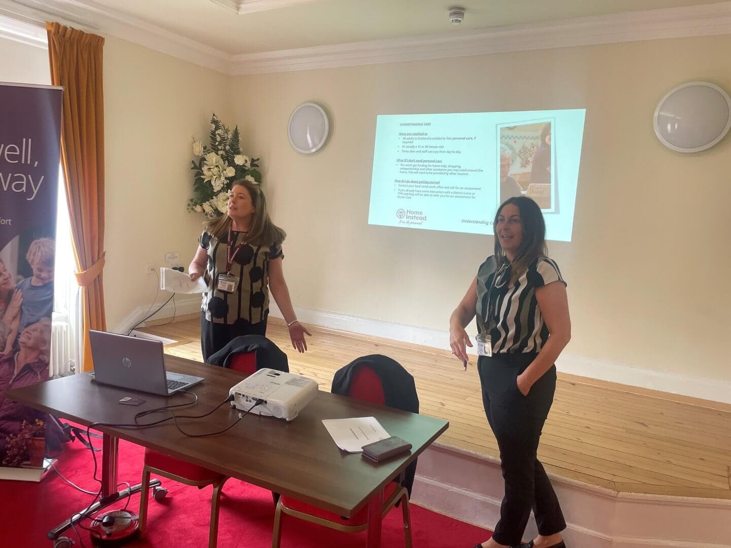 Two women giving a presentation in a room with a projection screen and a table with a laptop and documents. - Home Instead