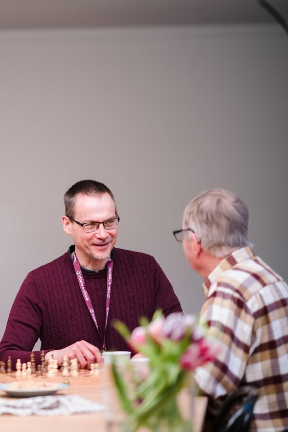 Two men sitting at a table with chess pieces, smiling and talking, with a vase of flowers in the foreground. - Home Instead