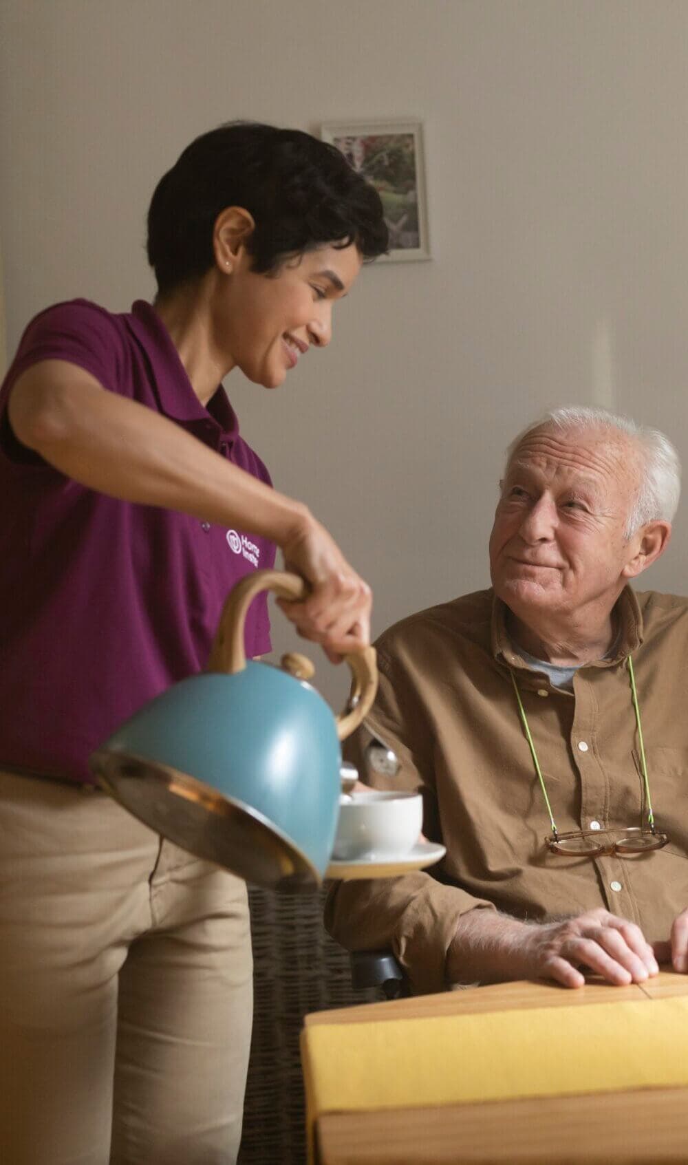 A Care Professional in a purple shirt pours tea for an elderly client, who is sitting and smiling at her. - Home Instead Bournemouth & Christchurch